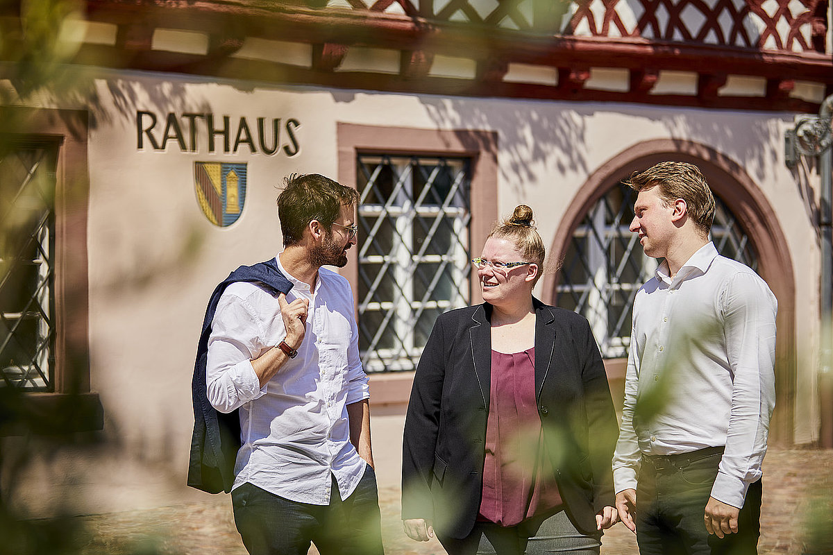 Zwei Männer und eine Frau stehen vor einem Rathaus und unterhalten sich